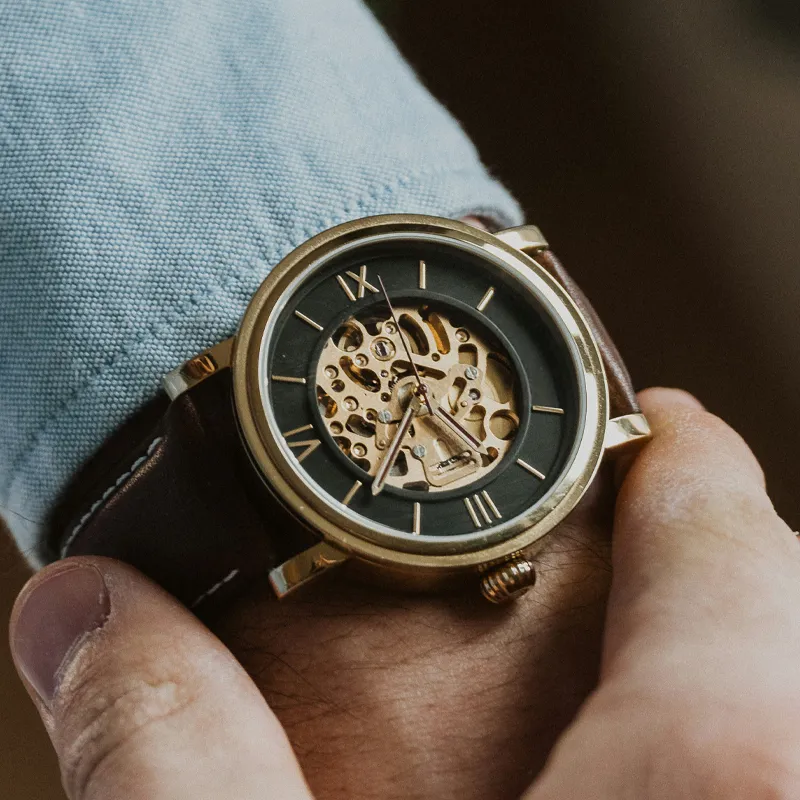 Close-up of a gold skeleton watch on a wrist, featuring a black dial and brown leather strap. Shop at Bryan Jewelry.