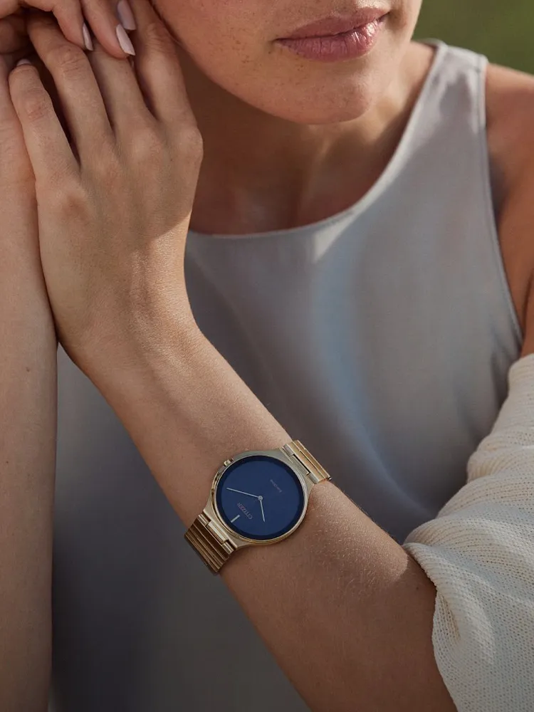 A woman wearing a gold watch with a blue face, resting her hand on her chin, showcasing a stylish accessory.