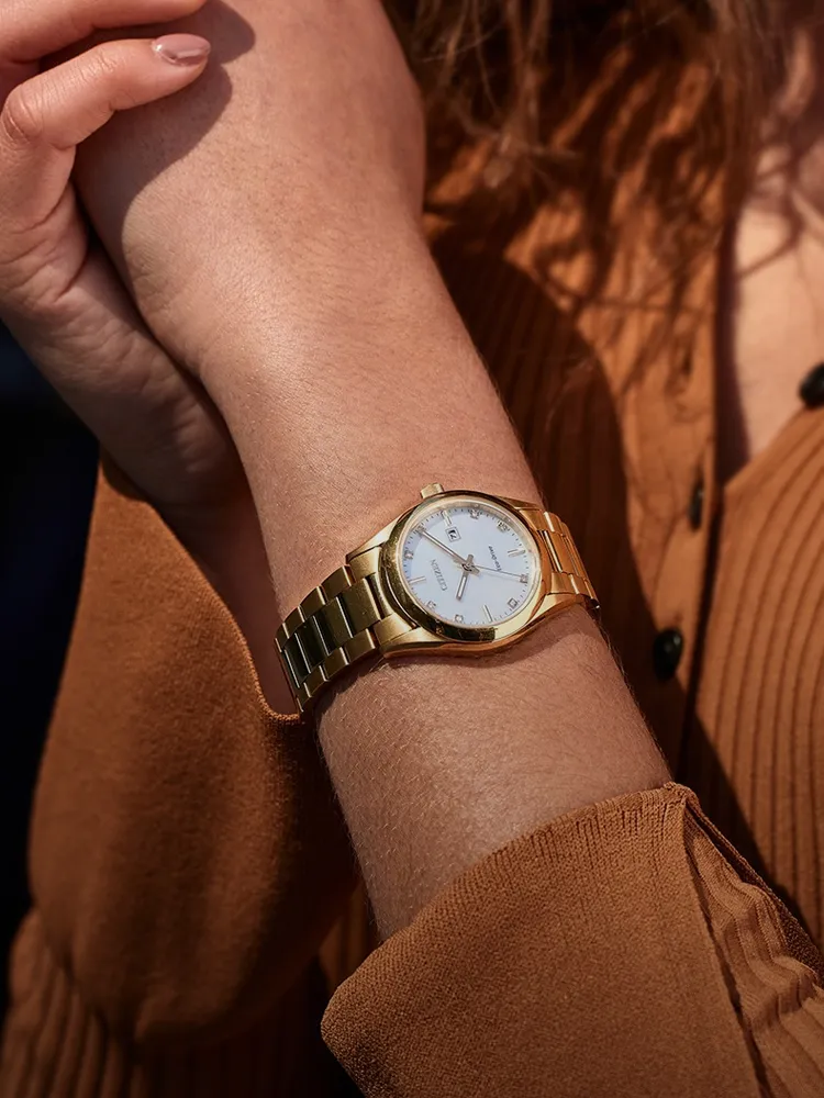 Gold women's watch with a white face, worn on a wrist, against a brown textured background. Shop at Bryan Jewelry.
