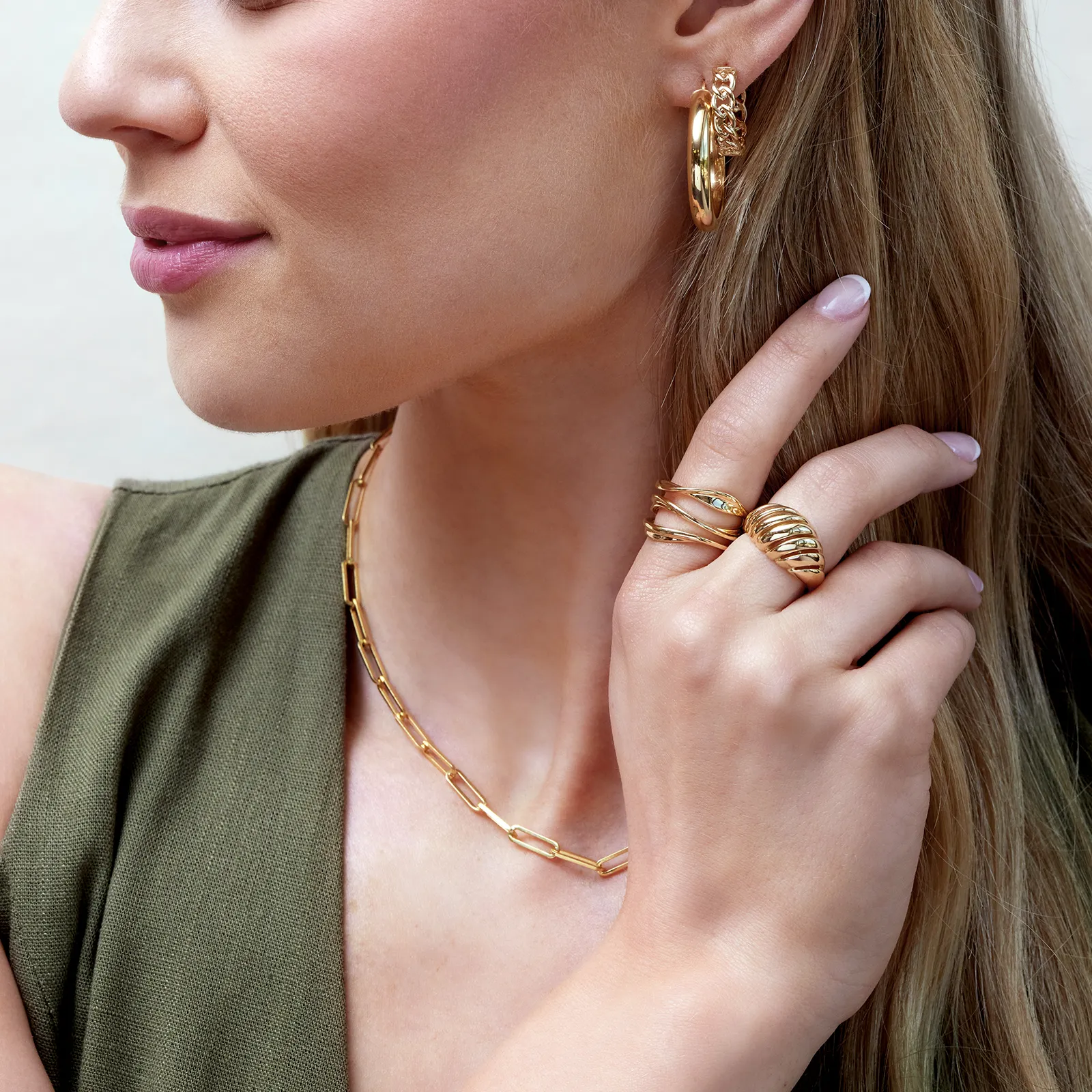 Woman wearing gold jewelry: hoop earrings, layered rings, and a chain necklace.