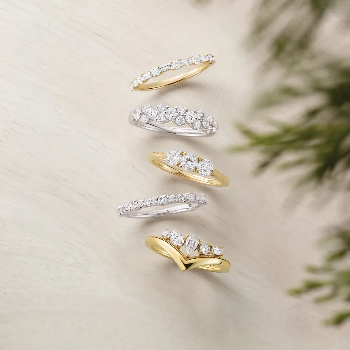 Five diamond rings in gold and silver displayed on a light surface with greenery.