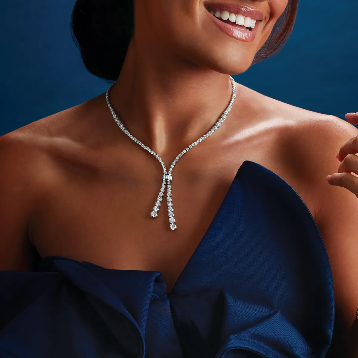 A woman wearing a diamond tennis necklace and a blue dress, smiling against a dark background.