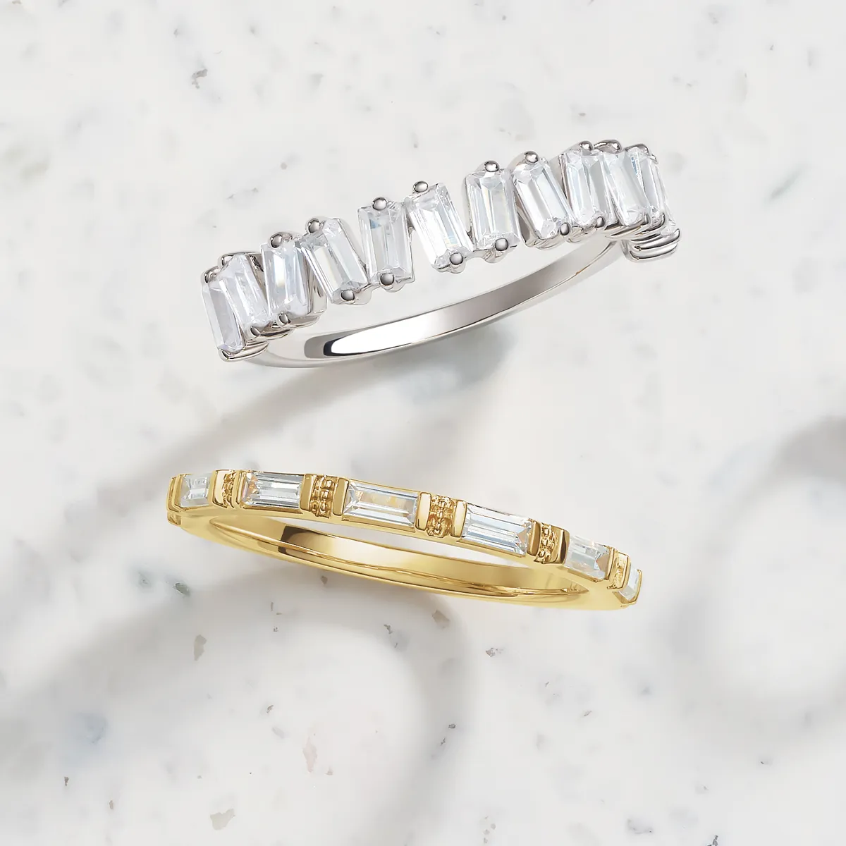 Two rings with baguette diamonds, one in silver and one in gold, on a marble surface.