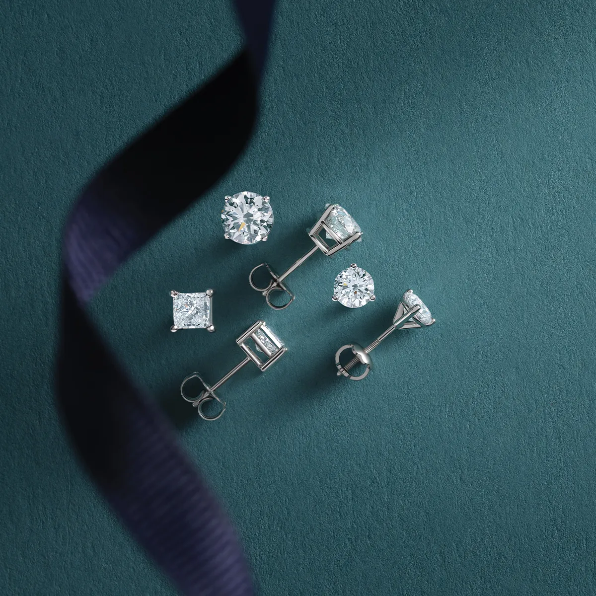 Diamond stud earrings on a teal background with a dark ribbon accent.