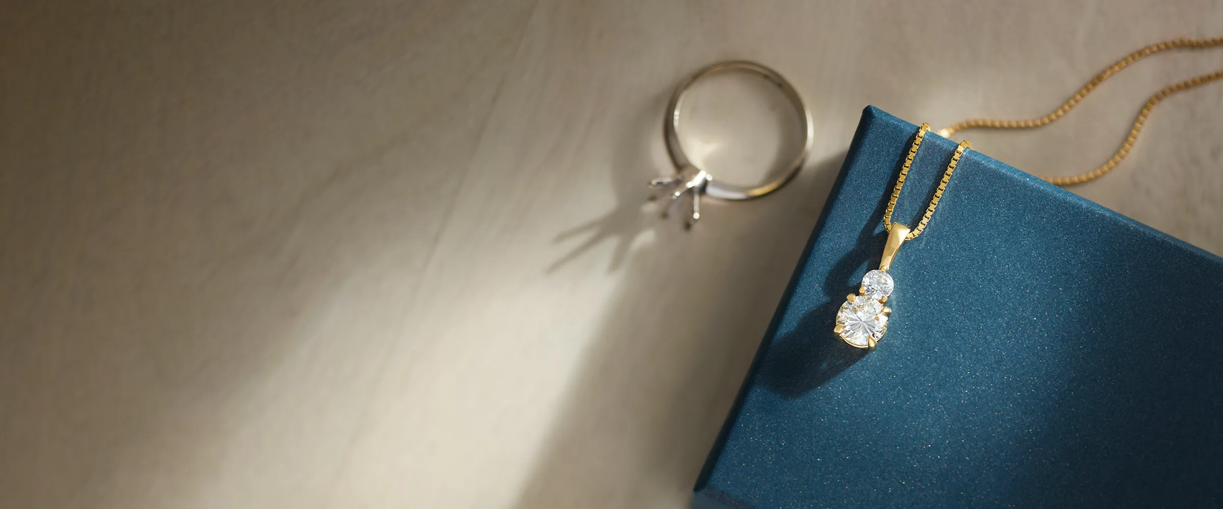 Gold pendant with diamonds on a blue box beside a ring setting on a light surface.