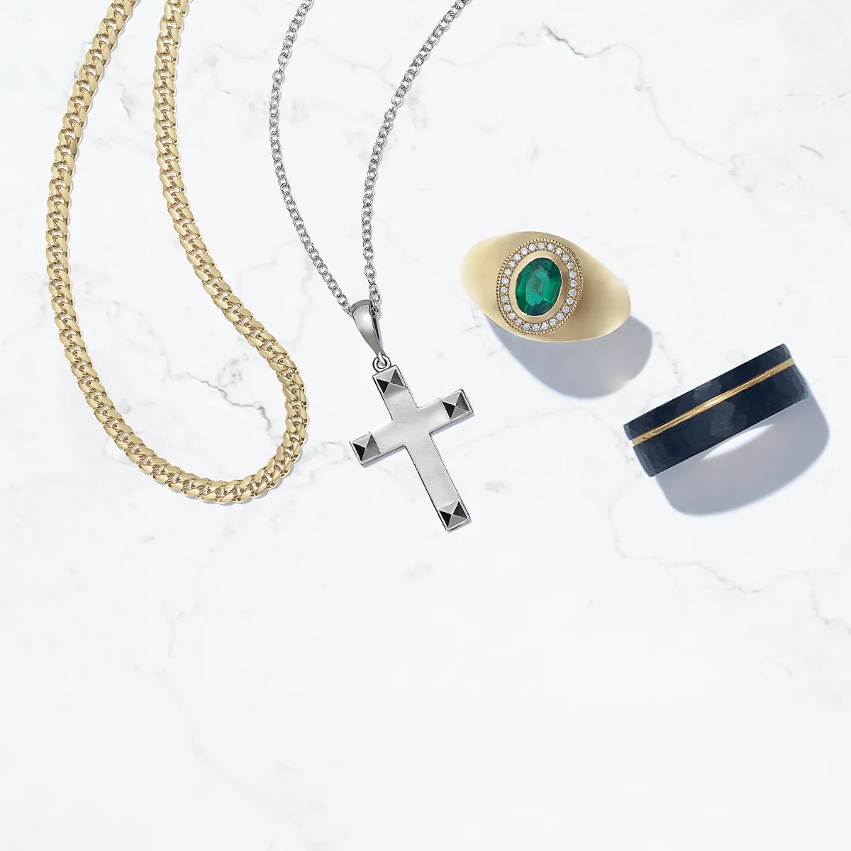 Gold chain, silver cross necklace, emerald ring, and black band on marble surface.