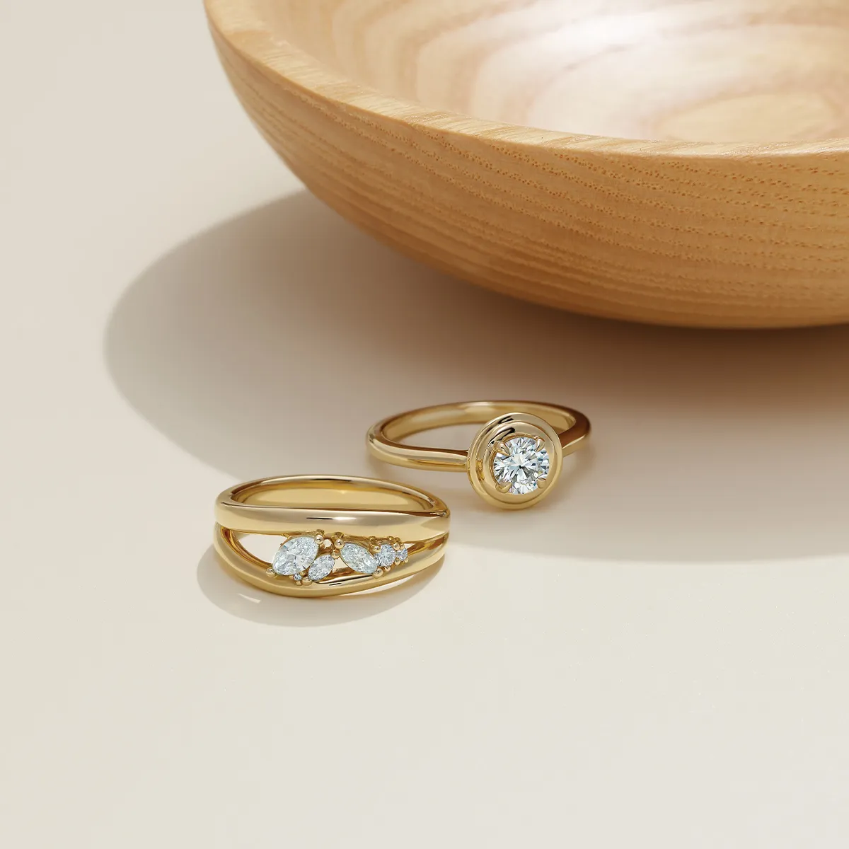 Two gold rings with diamonds displayed near a wooden bowl on a light surface.