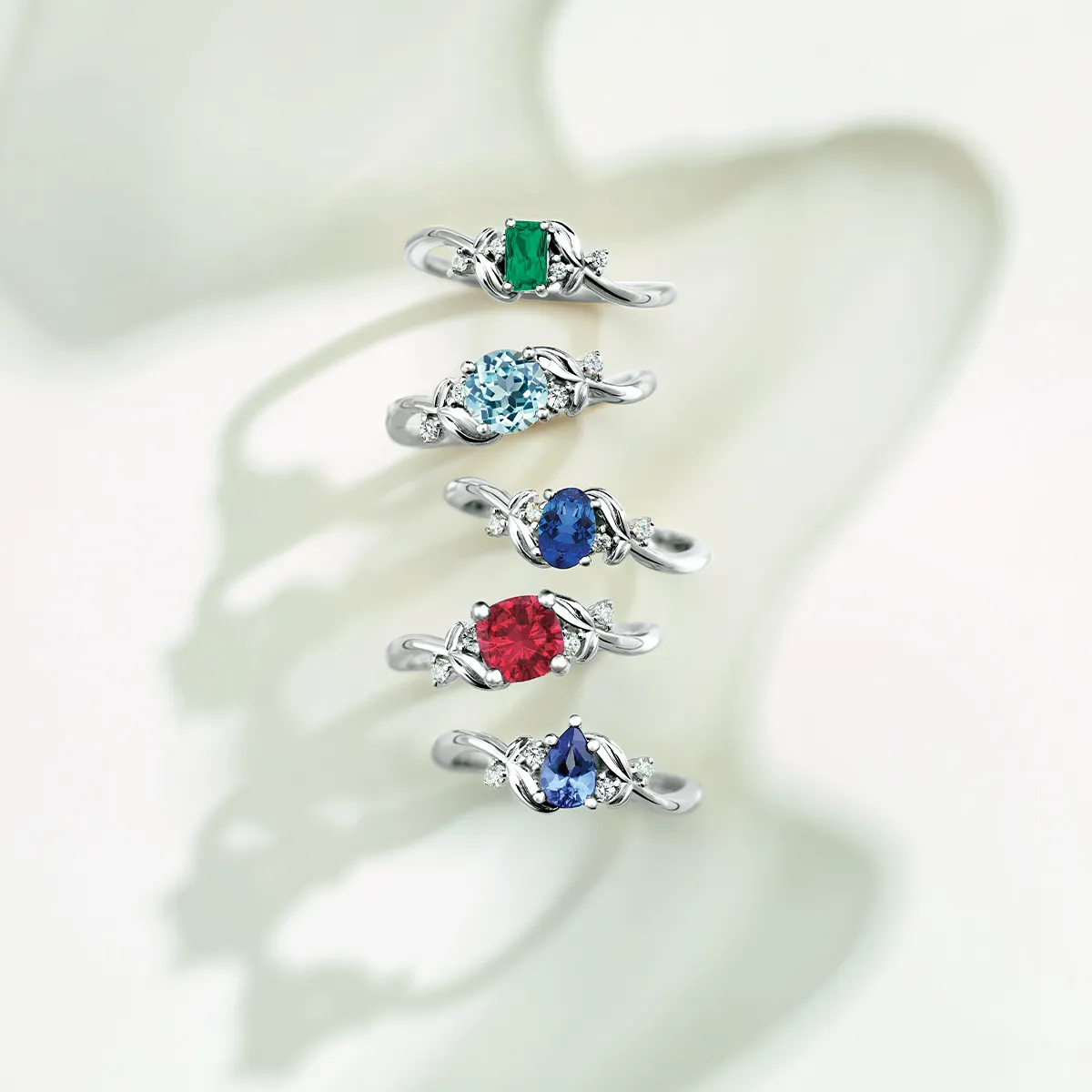 Five silver rings with colorful gemstones: green, light blue, blue, red, and dark blue, displayed on a light background.