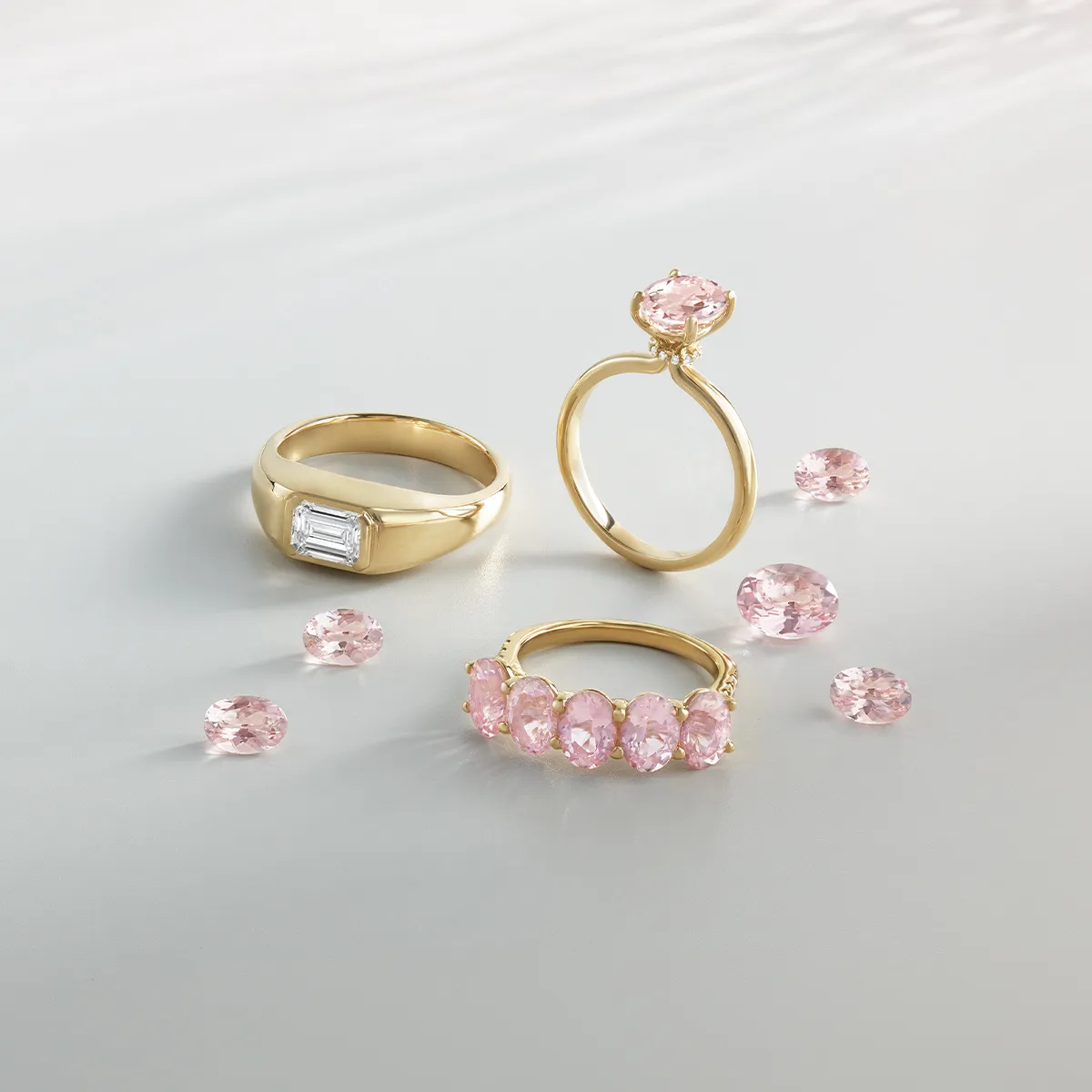 Three gold rings with pink gemstones and loose pink stones on a light surface.