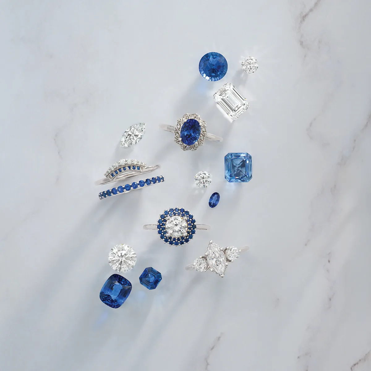 A collection of sapphire and diamond rings and loose gemstones on a marble surface.