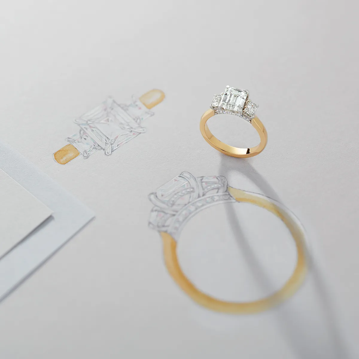 A gold engagement ring with a diamond next to its design sketch on paper.