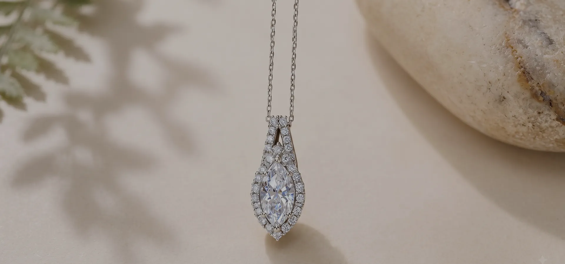 A pear-shaped diamond pendant with a halo on a delicate chain, set against a soft background with a stone and leaf shadow.