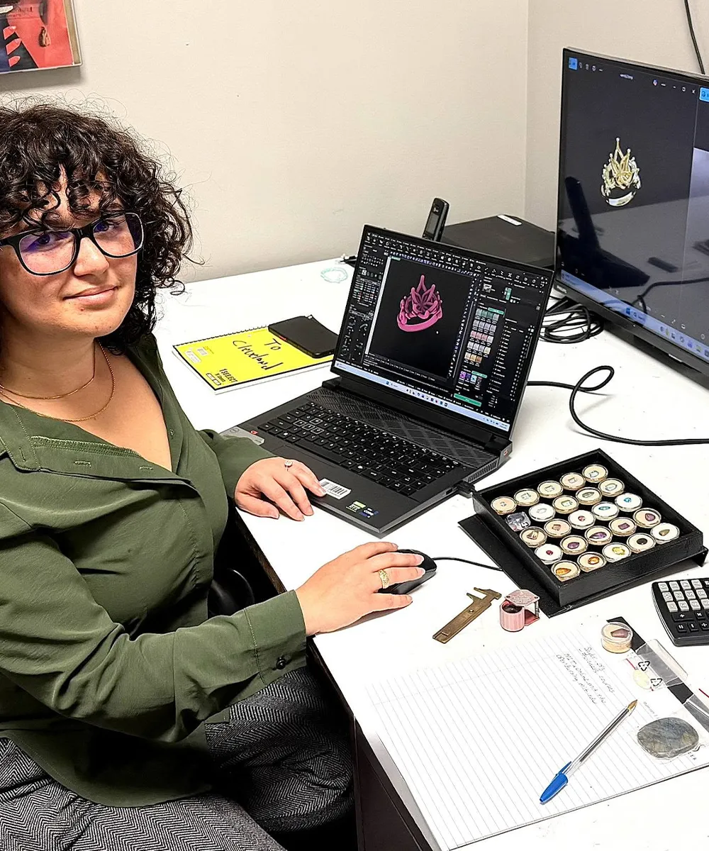 A person with curly hair works on jewelry design software at a desk with a laptop and monitor.