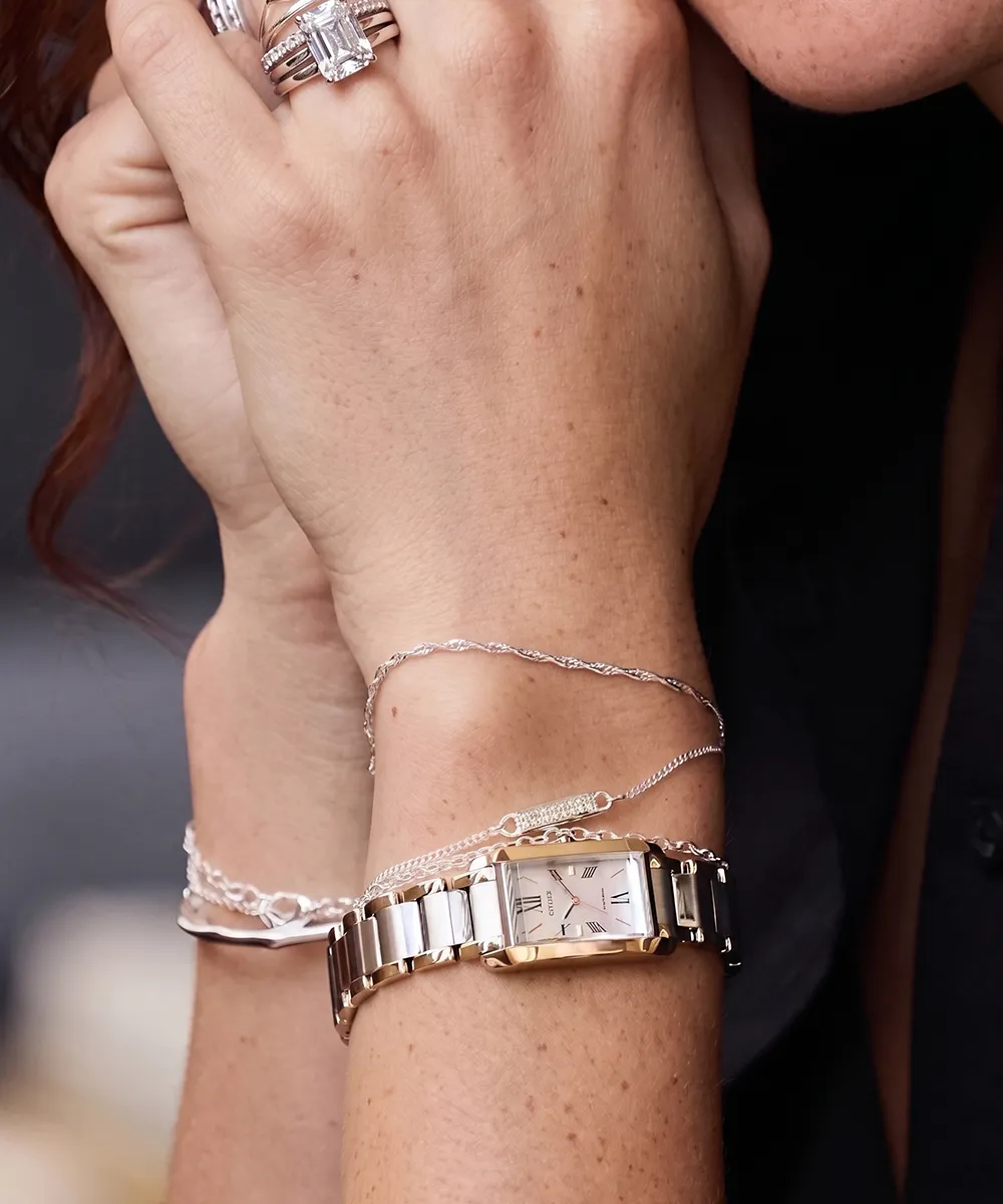 A close-up of a wrist wearing a silver watch, delicate bracelet, and rings with a large gemstone.