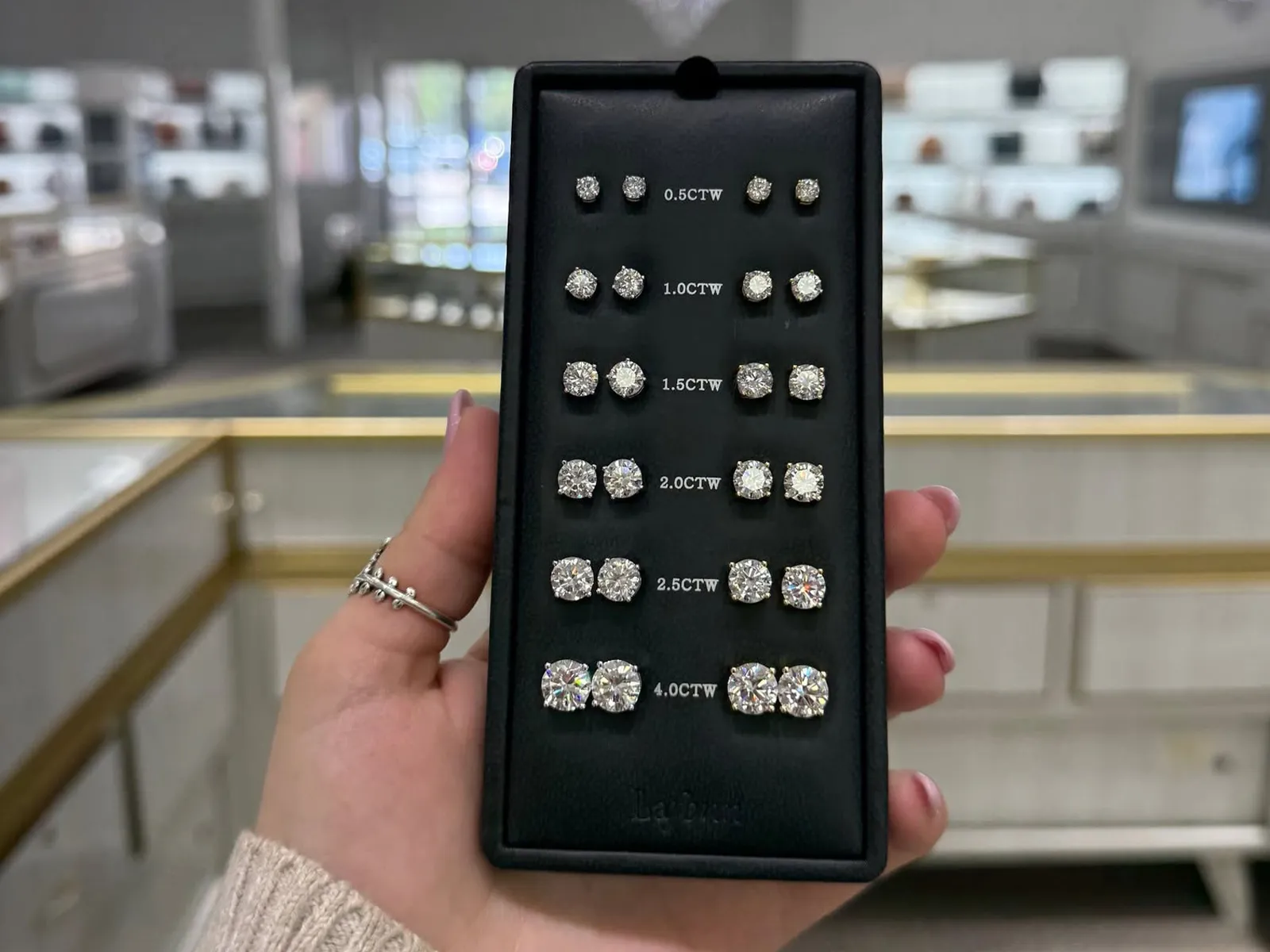 A hand holding a display of diamond stud earrings in various sizes, labeled by carat weight, inside a jewelry store.