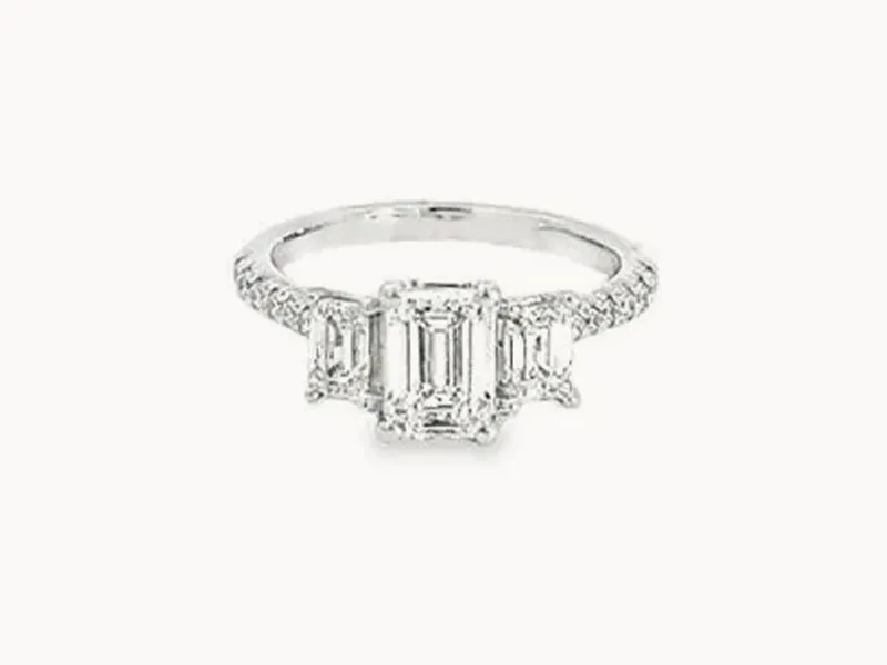 A three-stone diamond engagement ring with an emerald-cut center stone and side stones on a white band.