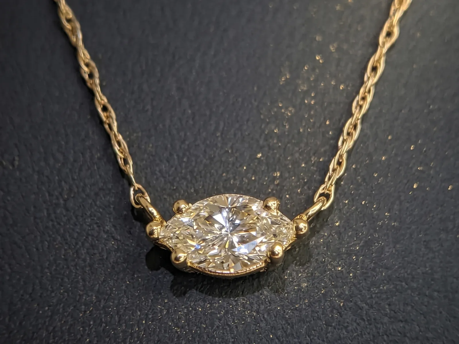 A gold necklace with a marquise-cut diamond pendant on a dark textured background.