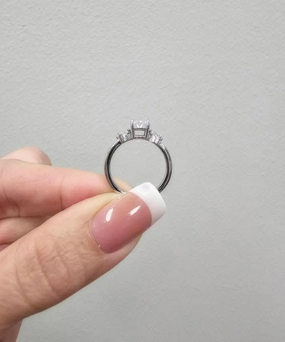A hand with a French manicure holds a diamond engagement ring against a plain background.