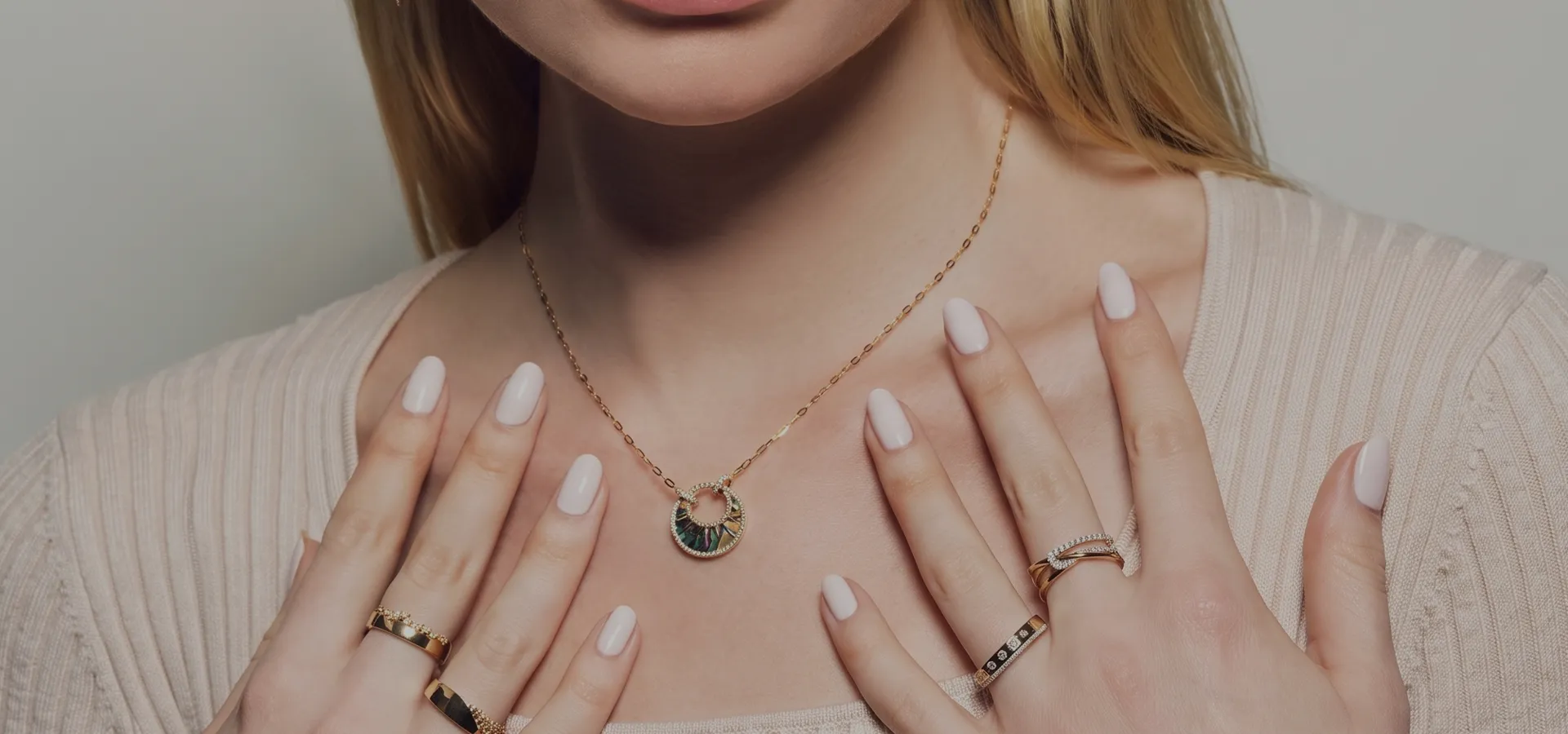 A woman wearing a gold necklace with a circular pendant, displaying manicured nails and multiple rings.