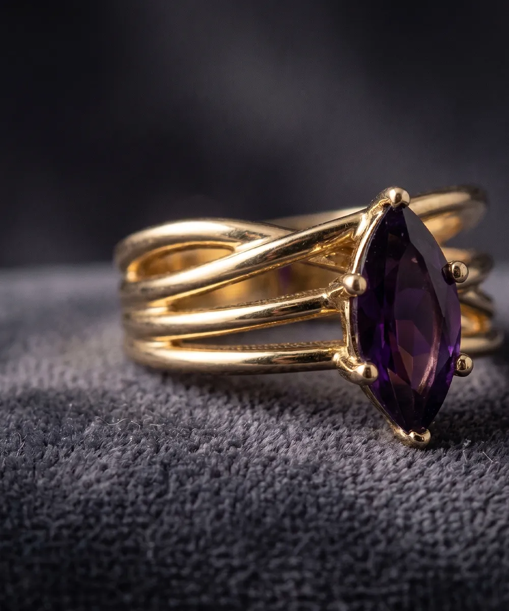 A gold ring with a marquise-cut purple gemstone on a textured gray surface.