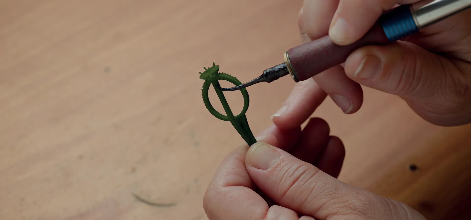 A person carving a green wax ring model with a tool on a wooden surface.