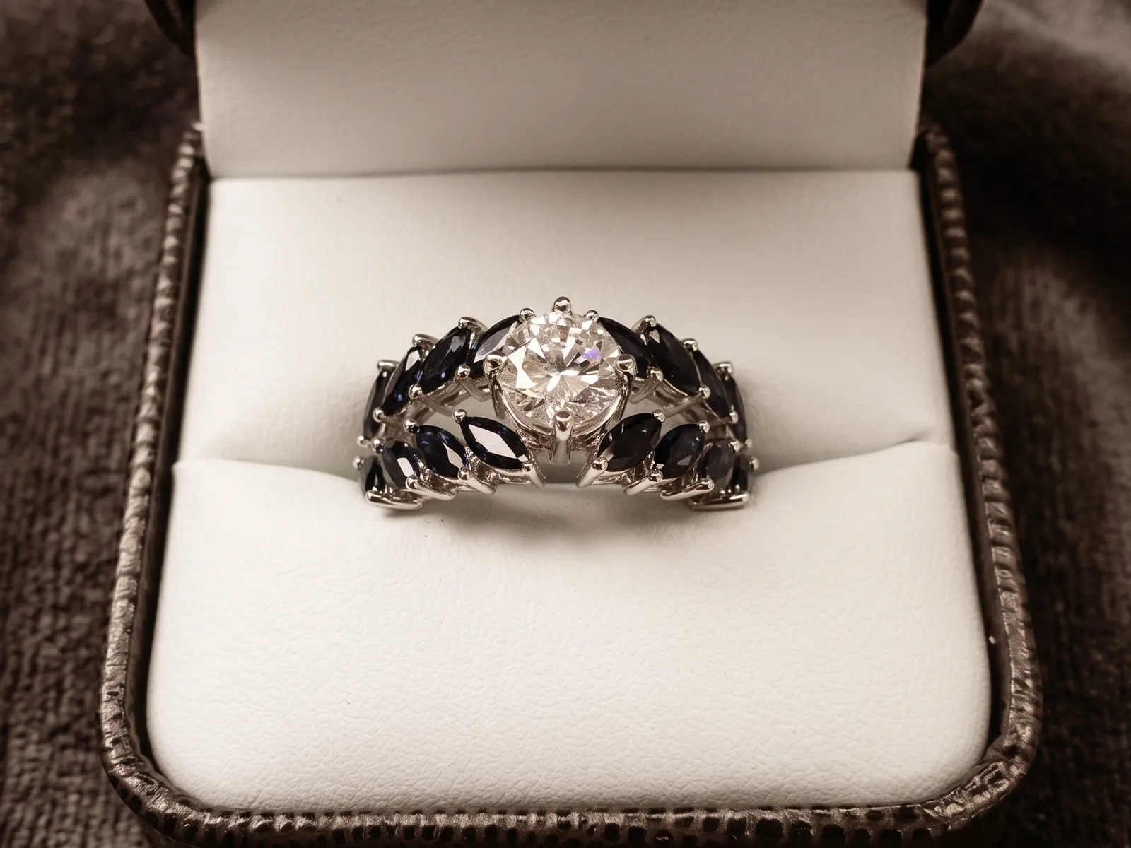 A silver ring with a central diamond and dark blue marquise stones in a jewelry box.
