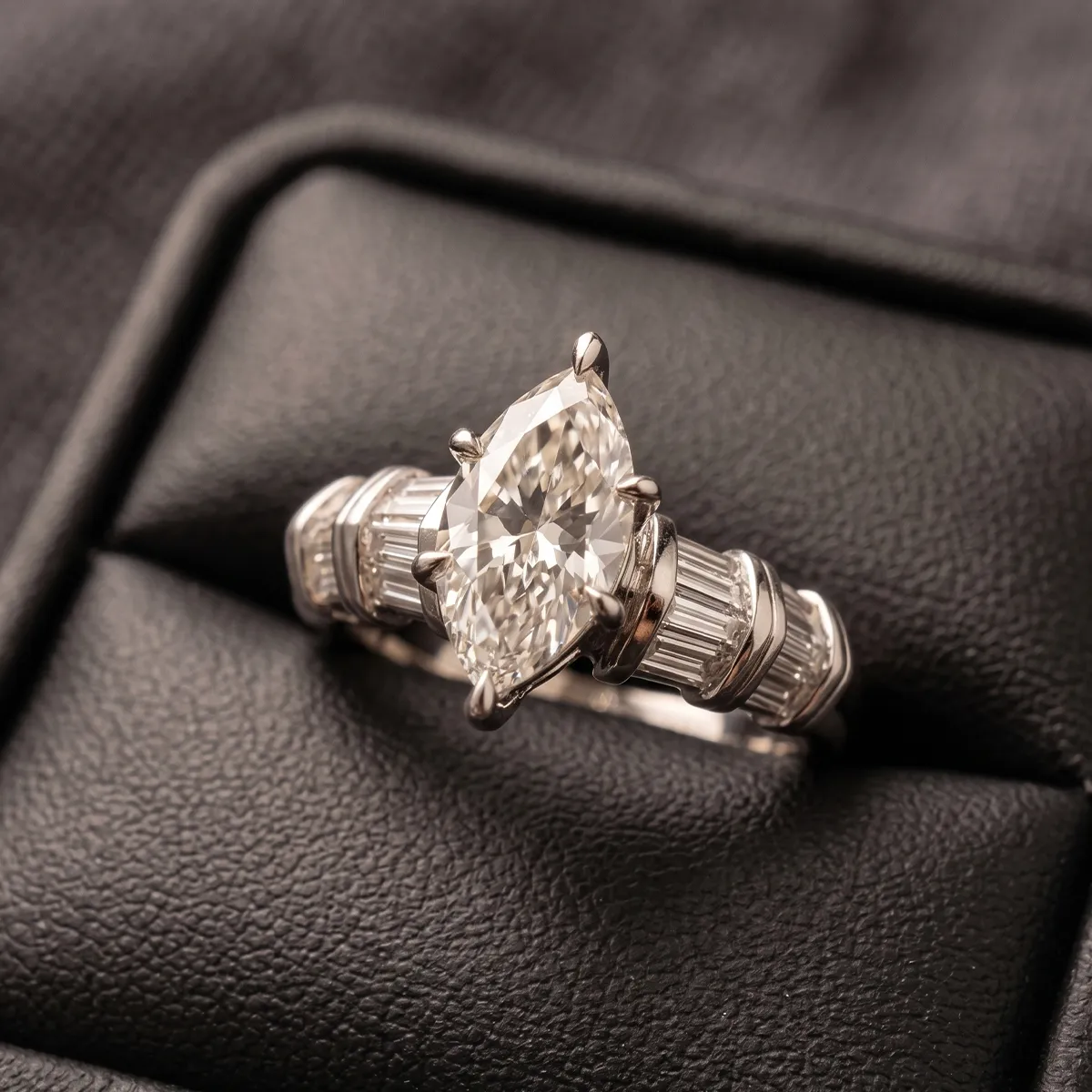 A marquise-cut diamond ring with baguette side stones on a black leather surface.