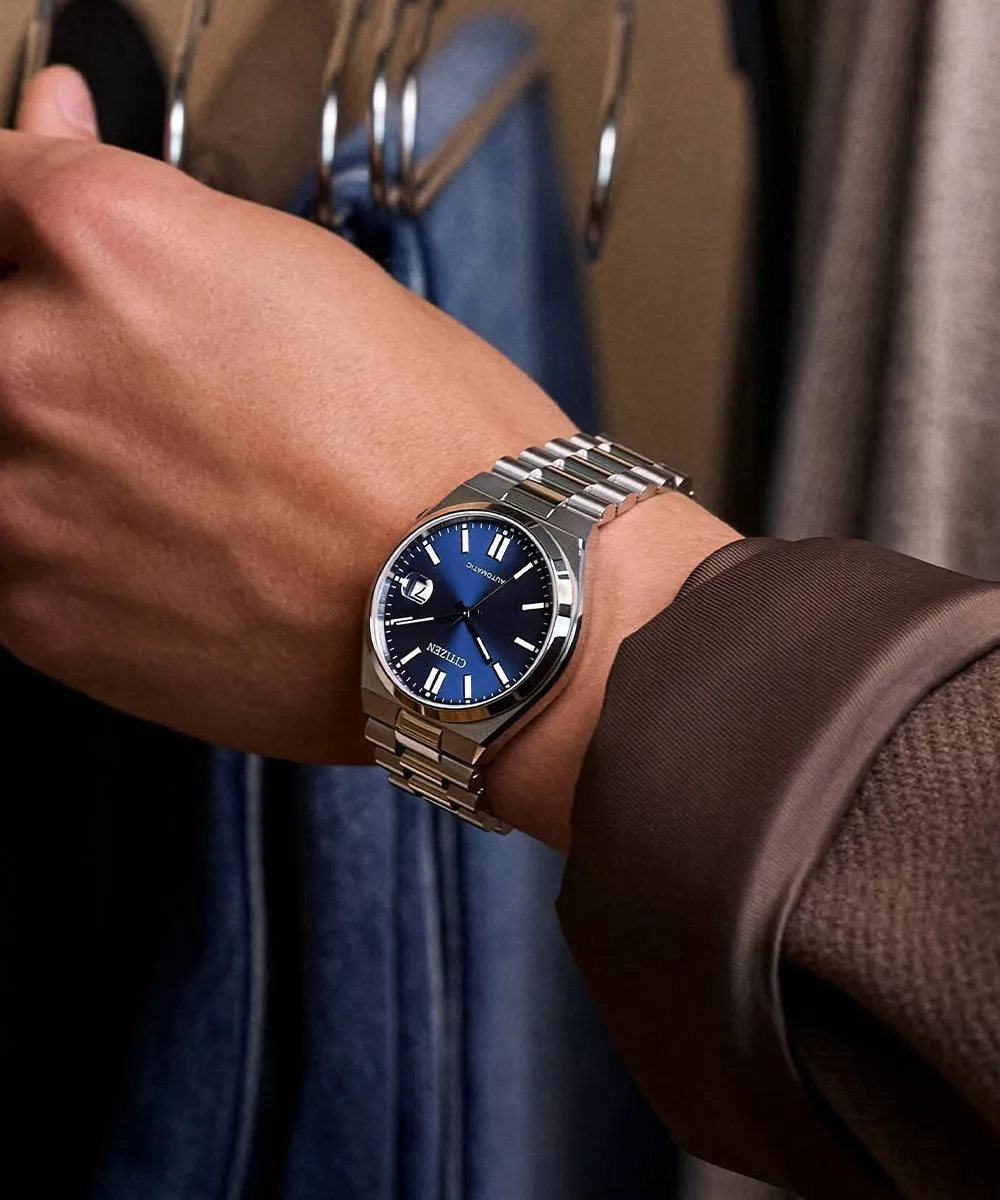 A person wearing a silver watch with a blue dial, standing near hanging clothes.
