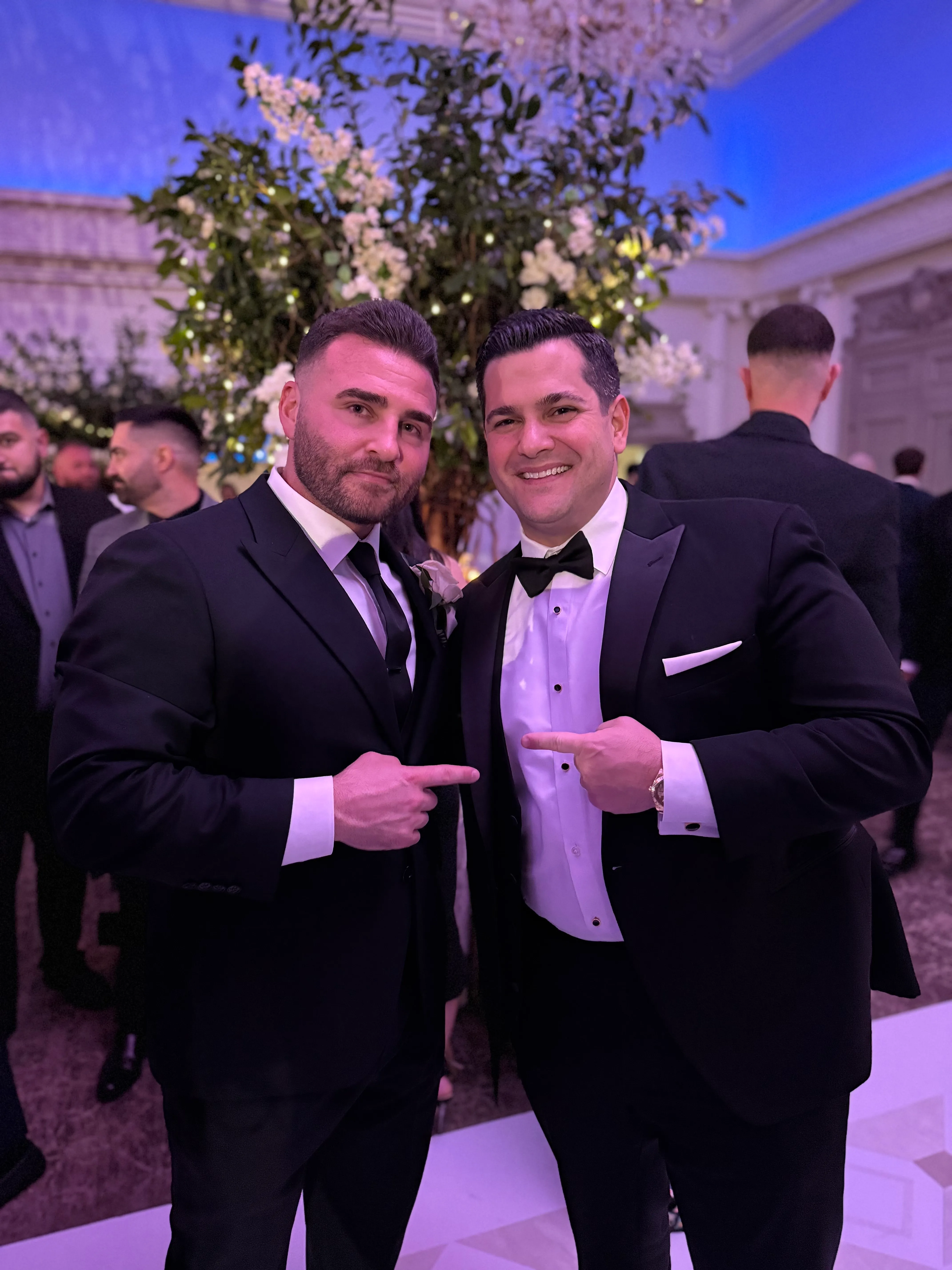 Two men in tuxedos posing together at a formal event, surrounded by elegant decor and guests in the background.
