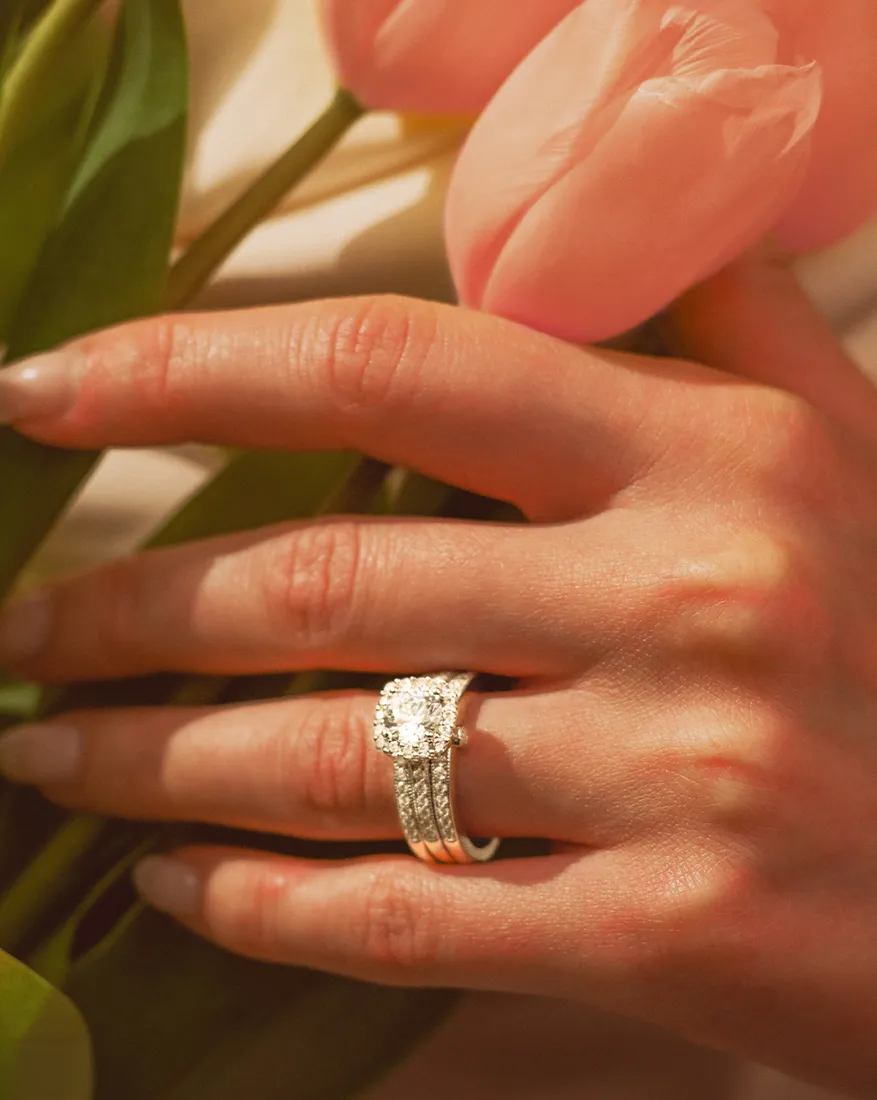 A hand with a diamond ring holds pink tulips. Visit Dahlkemper's Jewelry Connection for more details on engagement rings.