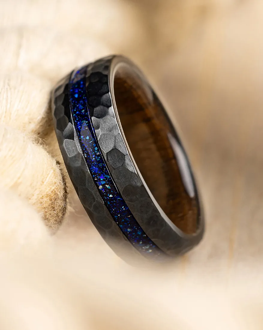 Hammered black ring with blue inlay on soft background. Click to explore more men's bands at Dahlkemper's Jewelry Connection.