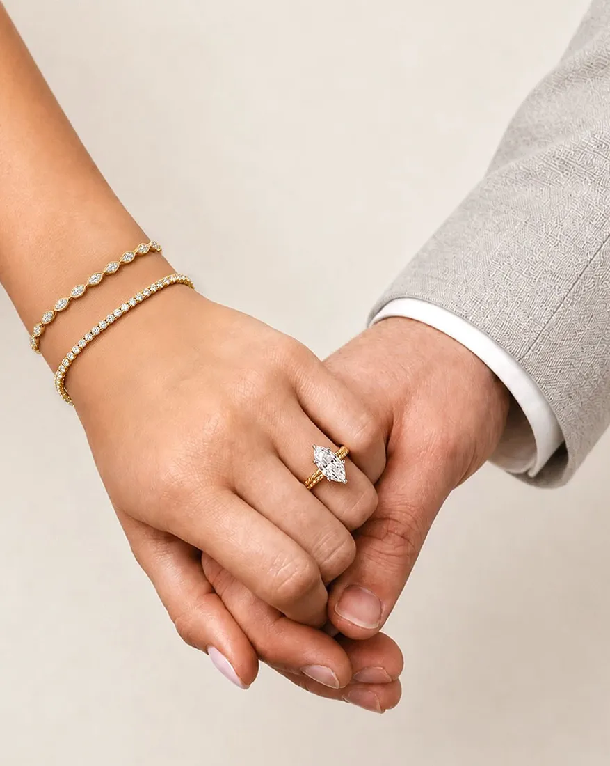 Two hands holding; one with a marquise diamond ring and gold bracelets, the other in a light suit.