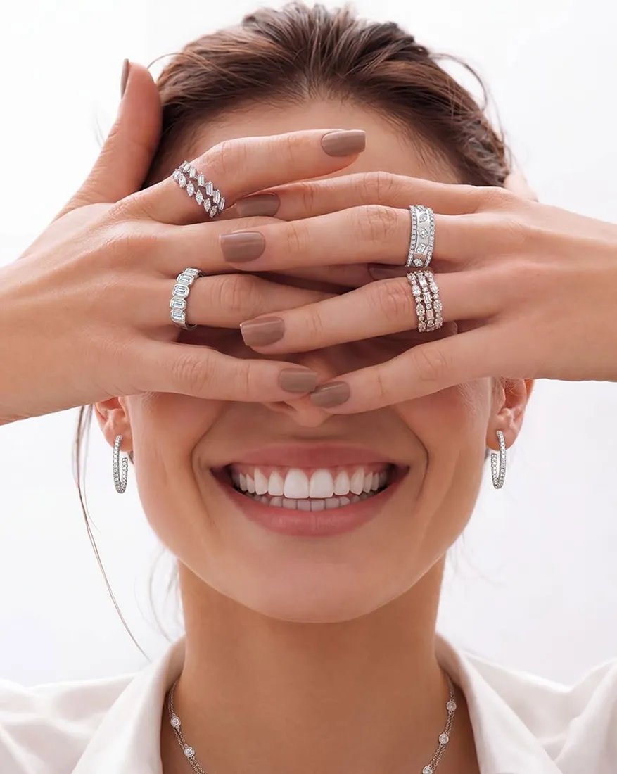 Smiling woman with hands covering eyes, wearing multiple silver rings and earrings.