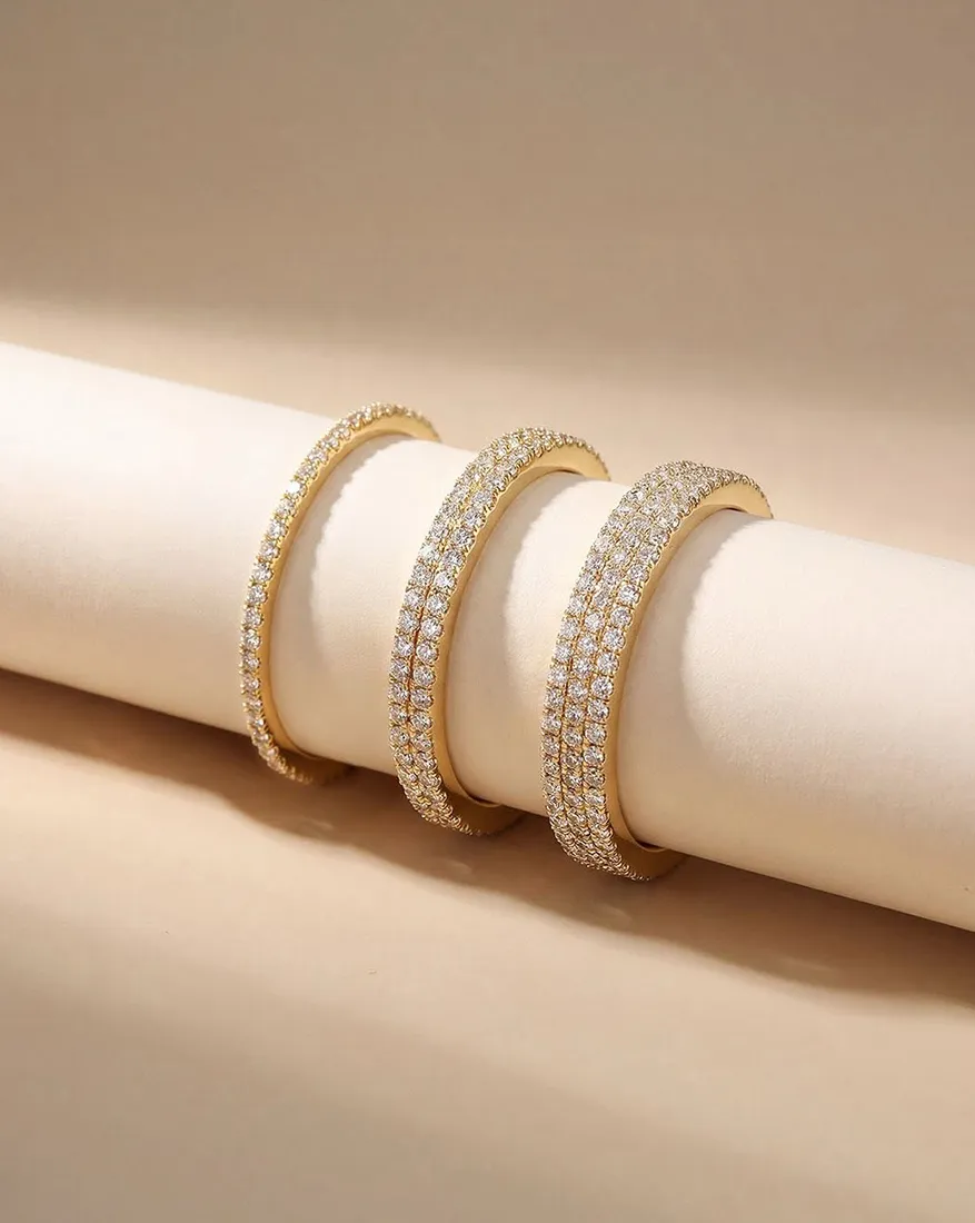 Three gold bangles adorned with rows of sparkling diamonds displayed on a beige background.