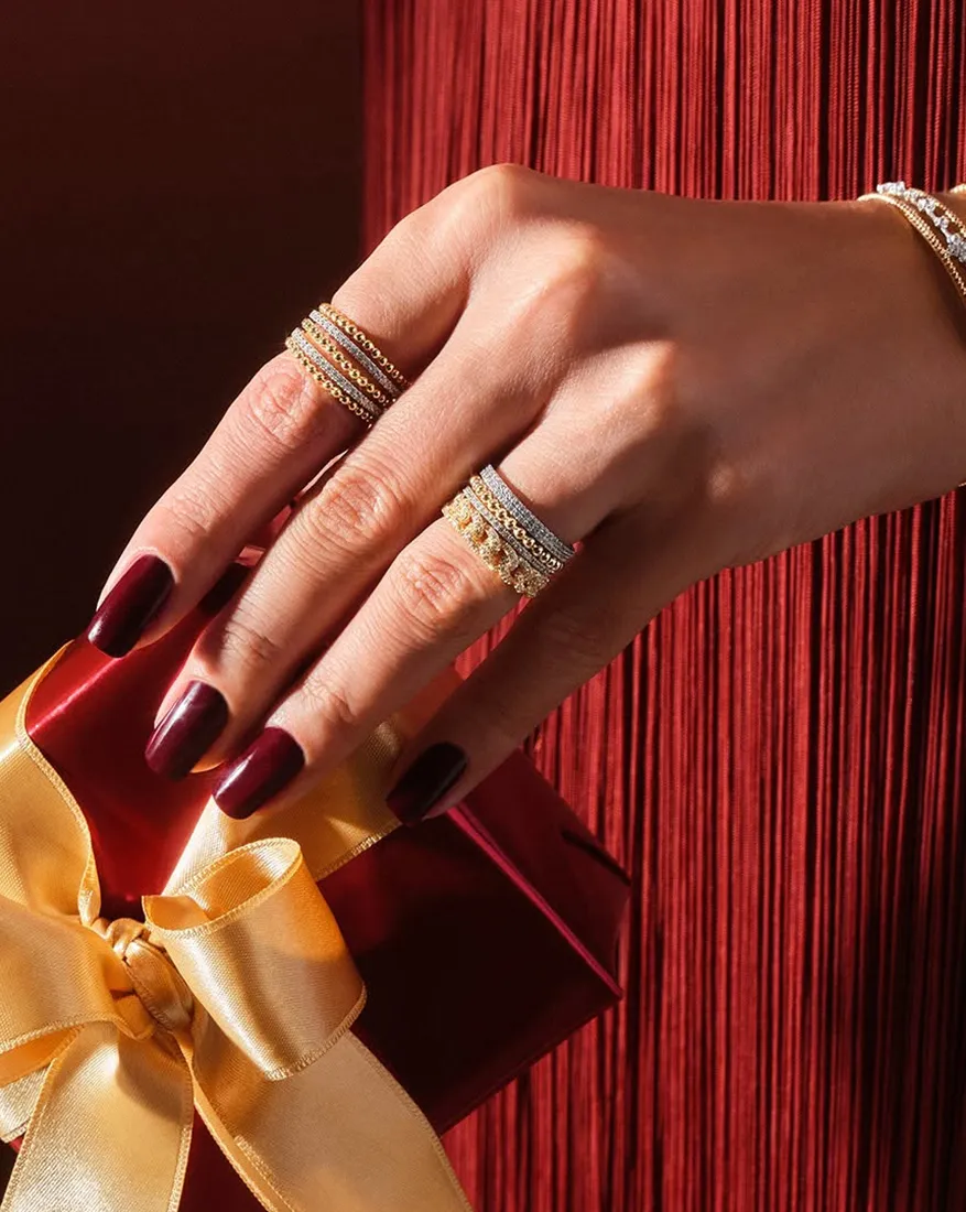 Hand with gold rings and dark nails holding a red gift box with a gold ribbon.