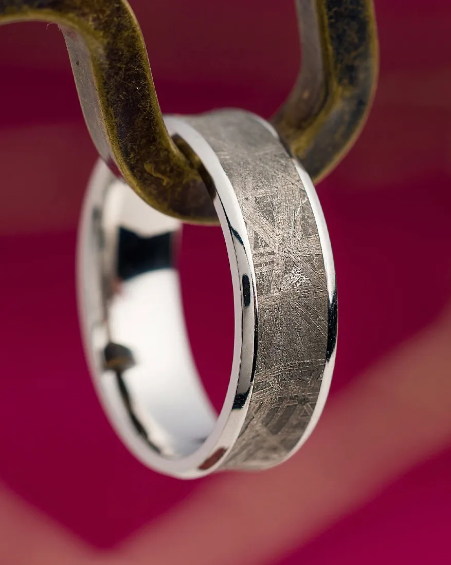 A silver ring with a textured surface hanging on a hook against a pink background.
