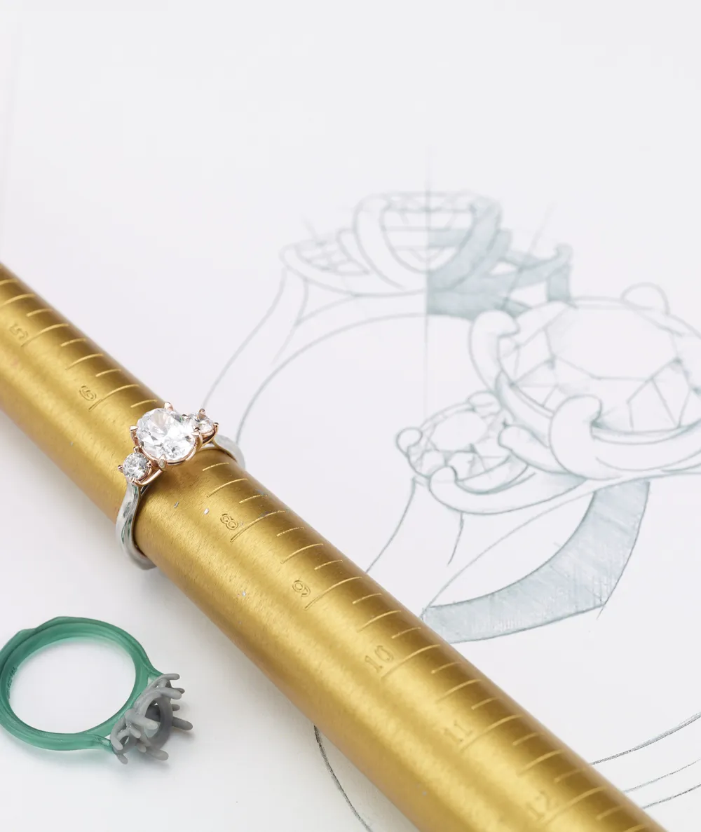 Engagement ring on a gold sizing mandrel with a sketch of a ring design in the background.