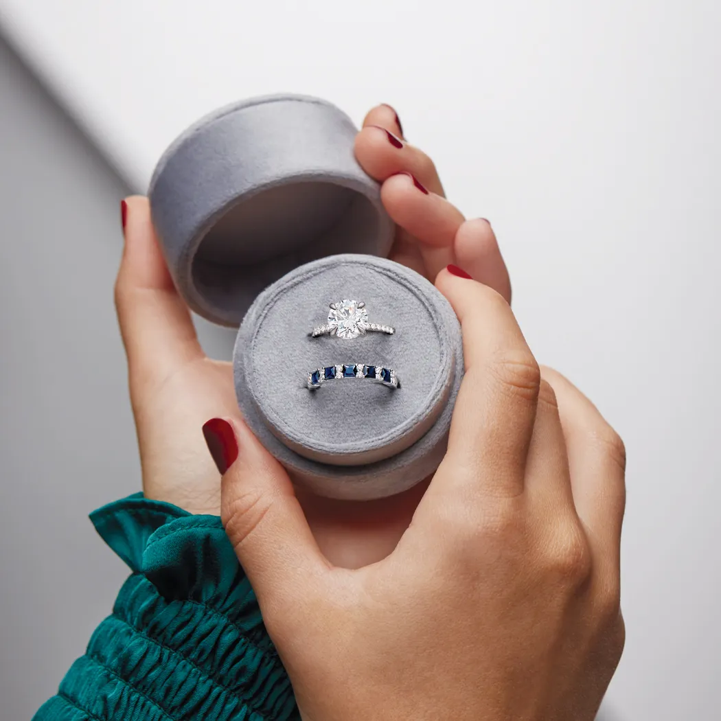 A hand holding a gray ring box containing a diamond engagement ring and a sapphire band.
