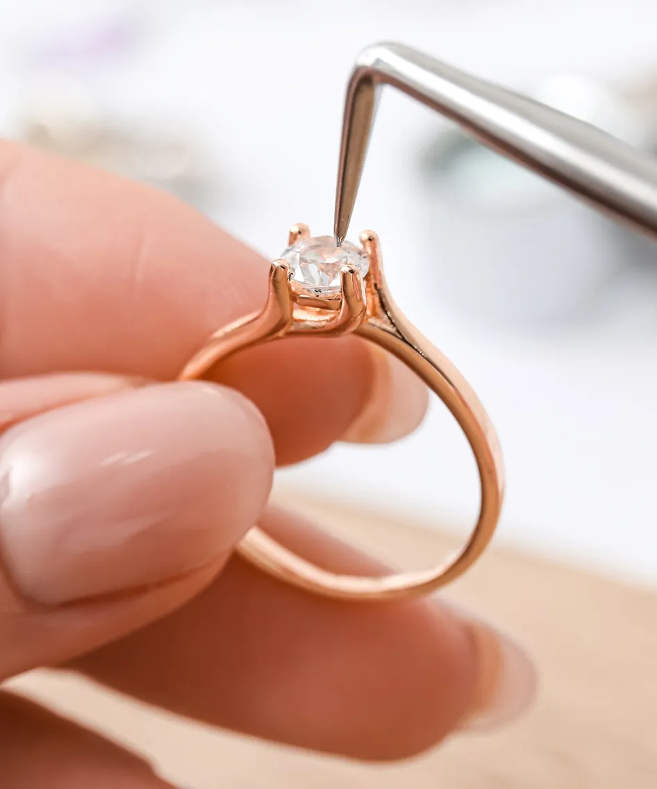 A hand holding a rose gold engagement ring with a diamond, as a tool adjusts the stone in its setting.