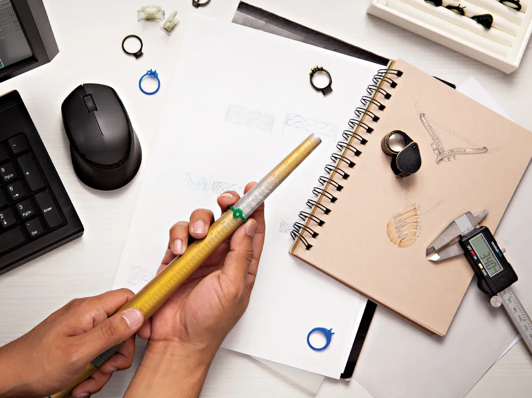 A hand holding a ring sizer tool over sketches and jewelry designs on a workspace with a computer and tools.