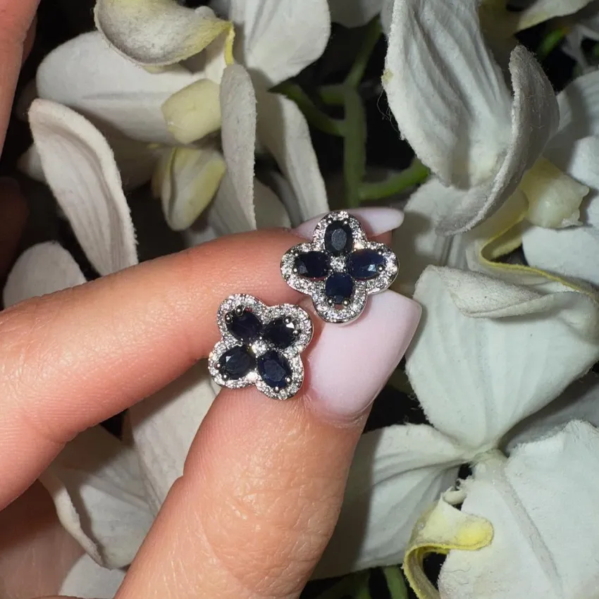 Sapphire flower earrings with diamond accents held between fingers, surrounded by white flowers.