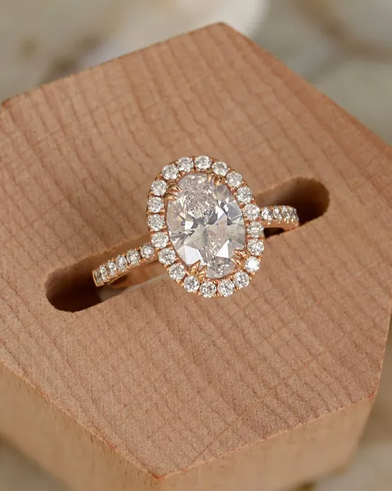 Engagement ring with an oval diamond surrounded by smaller diamonds, displayed on a wooden stand.