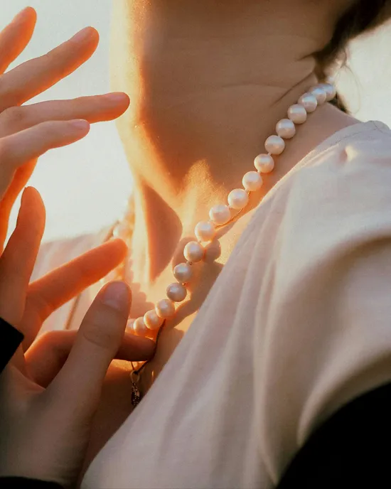 A close-up of a woman wearing a pearl necklace, gently touching it with her fingers in soft lighting.