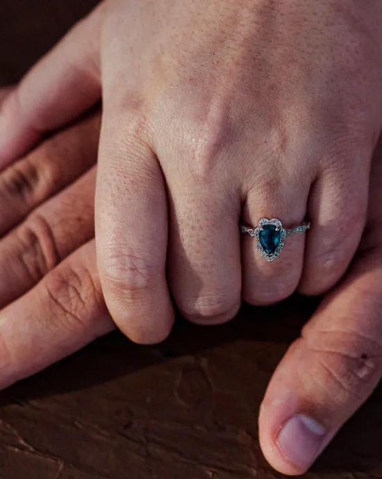 A close-up of a hand wearing a heart-shaped sapphire ring, resting on another hand.