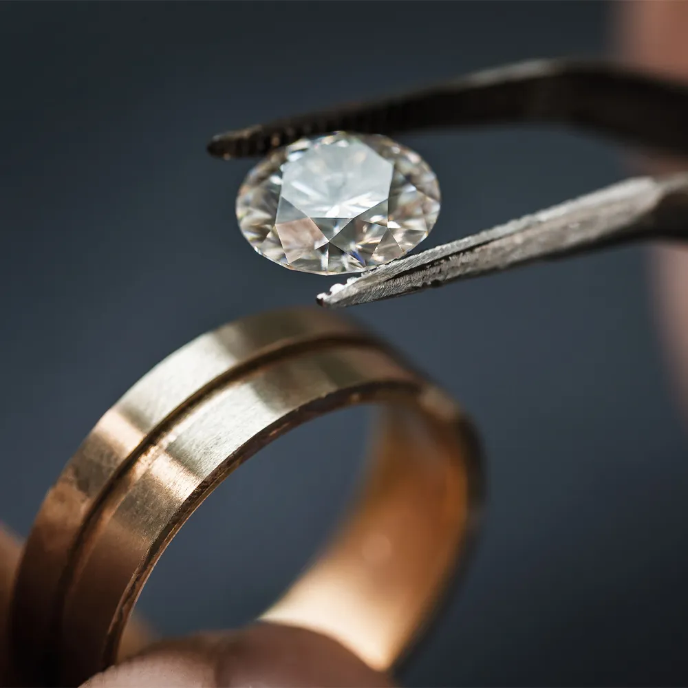 A jeweler using tweezers to hold a diamond above a gold ring, showcasing fine jewelry craftsmanship.