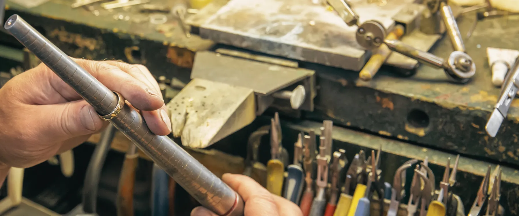 A jeweler's hands working on a metal piece at a cluttered workbench, showcasing craftsmanship. Click to contact Dickinson Jewele