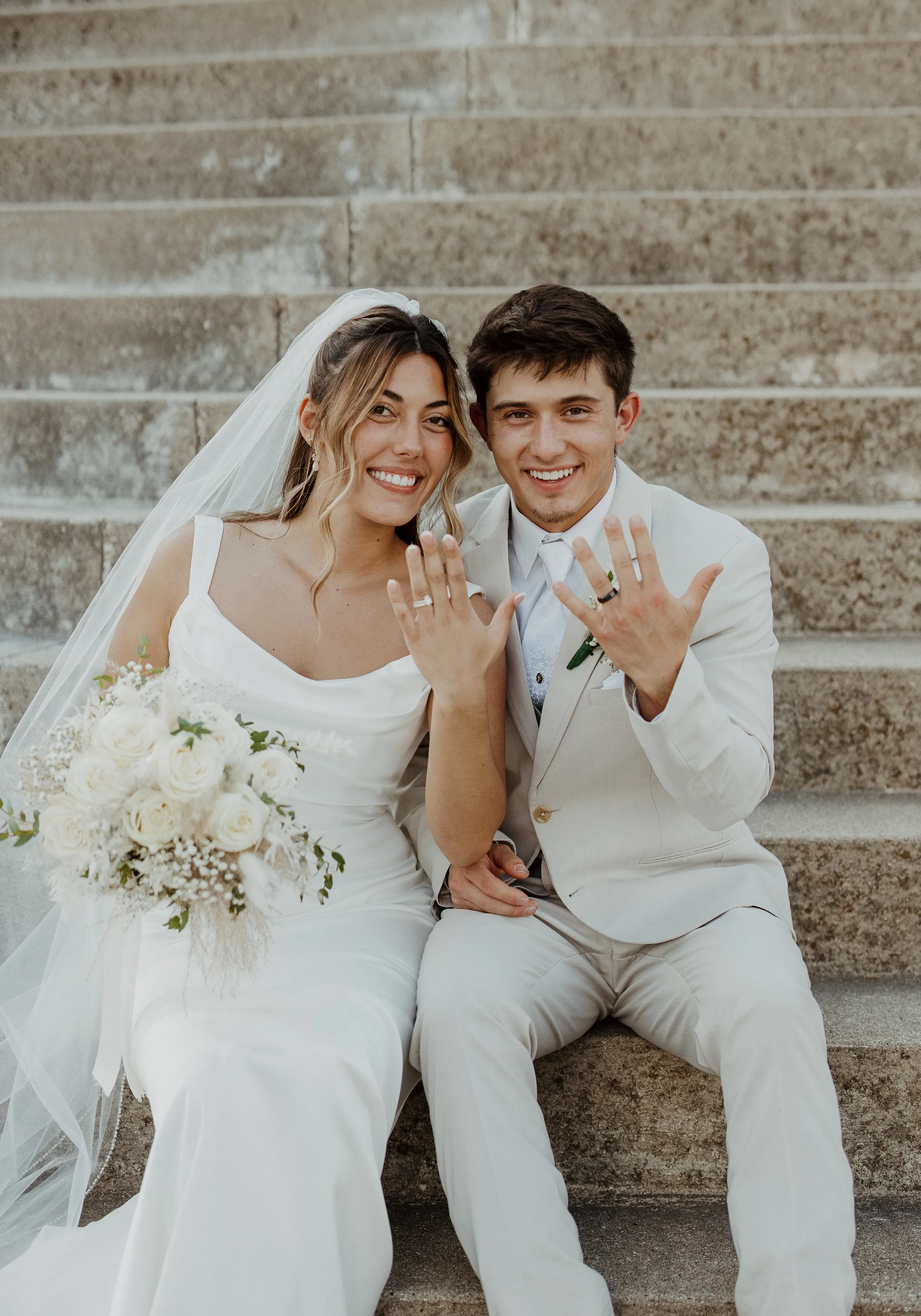 Bride and groom smiling outdoors with autumn trees in the background.