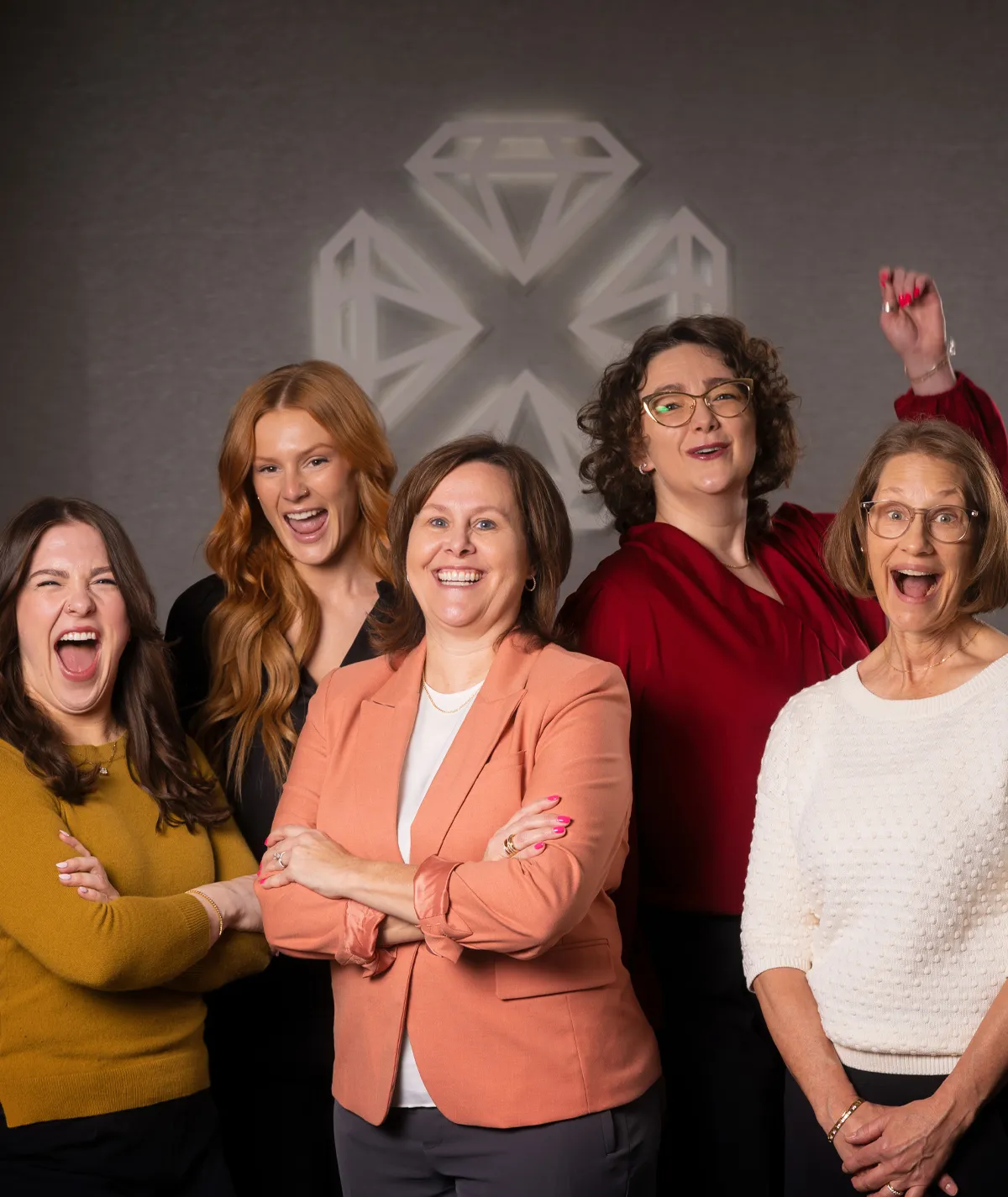 Five women smiling, standing together in front of a diamond logo at Diedrich Jewelers.