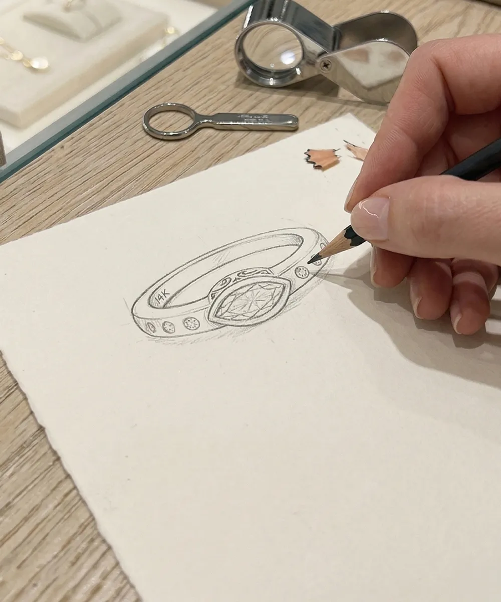 A hand sketching a detailed ring design on paper with jewelry tools nearby.