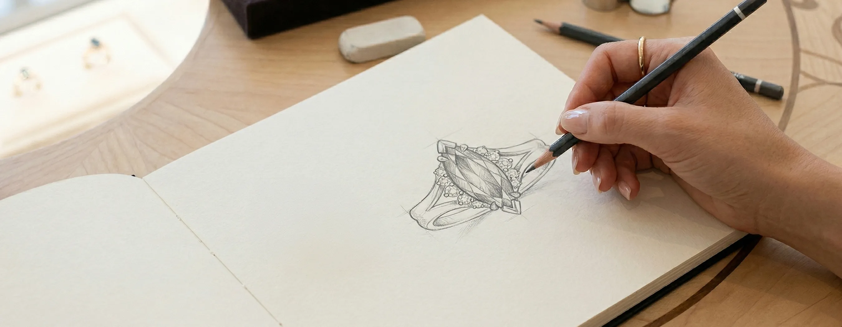 Hand sketching an intricate ring design on paper with pencils nearby.